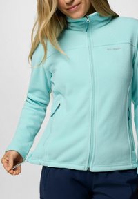 Giacca in pile azzurro chiaro con colletto alto, chiusura con zip e tasche laterali. Presenta un cordoncino regolabile all'orlo e una texture morbida.