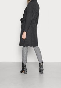 Tummanharmaa villasekoitetakki, jossa vyötäröllä vyö, yhdistetty harmaisiin ruudullisiin housuihin ja mustiin käärmekuvioituihin korkokenkiin.