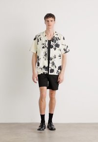Camicia a maniche corte floreale color crema con design nero, abbinata a pantaloni corti neri e scarpe nere; il modello è posizionato su uno sfondo neutro.