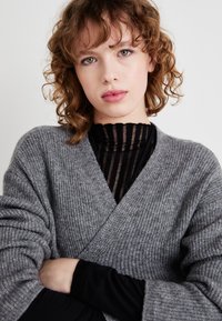 Grå strikket cardigan med ribbestruktur, med en V-halsdesign. Brukt over en svart gjennomsiktig stripete topp. Modell med krøllete hår.