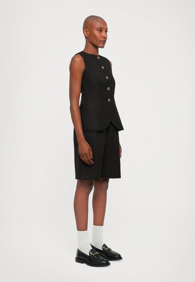 Black linen sleeveless vest with five button closure, paired with black shorts. Accessories include black loafers and white socks.