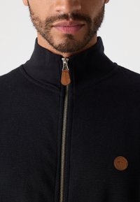 Hombre con barba corta que lleva un suéter negro con cremallera y tirador de cremallera marrón, además de un pequeño parche de cuero redondo de la marca en el pecho.