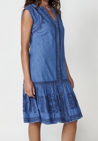 Robe en denim avec une fermeture à boutons sur le devant, des manches courtes et une jupe froncée. Elle présente un broderie bleu foncé et une forme décontractée en A.