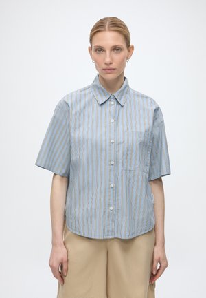 Mujer con cabello rubio que lleva una camisa de botones de manga corta a rayas azules y pantalones beige, de pie frente a un fondo blanco liso.