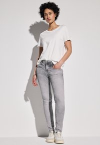 Jeans skinny grises con un acabado deslavado, combinados con una camiseta blanca sencilla. El modelo lleva zapatillas blancas y tiene una pose relajada.