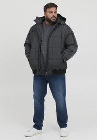 Svart boblejakke med hette, med en quiltet design, brede ermer og glidelås. Brukt over en grå hettegenser og parret med blå jeans.