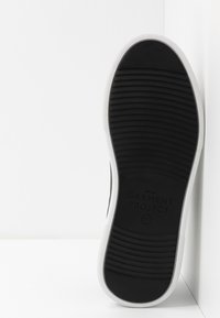 Svart gummisula med texturerad mönster. Vit övre kant och sida, med "GARMENT PROJECT" logotyp embärad på sidan.