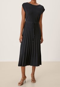 Donna che indossa un abito midi plettato senza maniche di colore nero con una cintura in vita annodata e sandali con tacco beige su un pavimento beige.