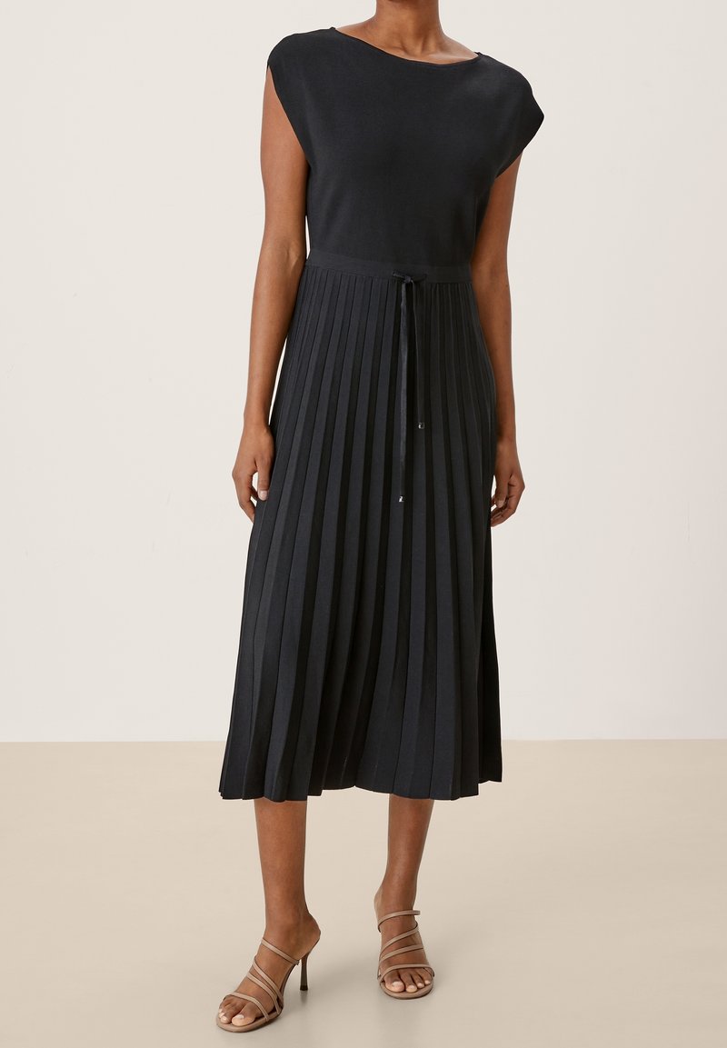 Donna che indossa un abito midi plettato senza maniche di colore nero con una cintura in vita annodata e sandali con tacco beige su un pavimento beige.