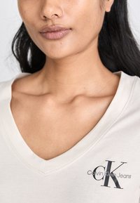 Šviesiai smėlinis V formos kaklo marškinėliai, pagaminti iš minkštos medvilnės, su tamsiai pilku logotipu "Calvin Klein Jeans" apačioje kairėje pusėje.