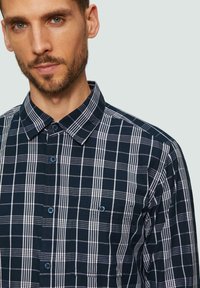 Marineblauw en witte geruite shirt met een button-down kraag, lange mouwen en een enkele borstzak met een blauwe knoopdetail.