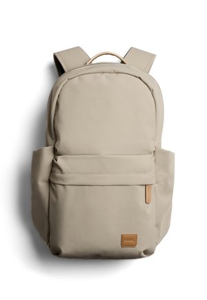 Zaino beige con spallacci imbottiti, tasca frontale con cerniera, tasche laterali e una piccola etichetta del marchio in pelle marrone in basso a destra.