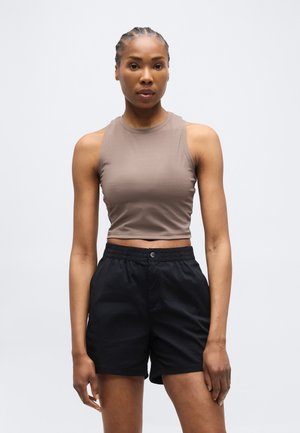 Fonott hajú nő, aki ujjatlan taupe színű rövid felsőt és fekete, magas derekú rövidnadrágot visel, egyszerű világos háttér előtt áll.