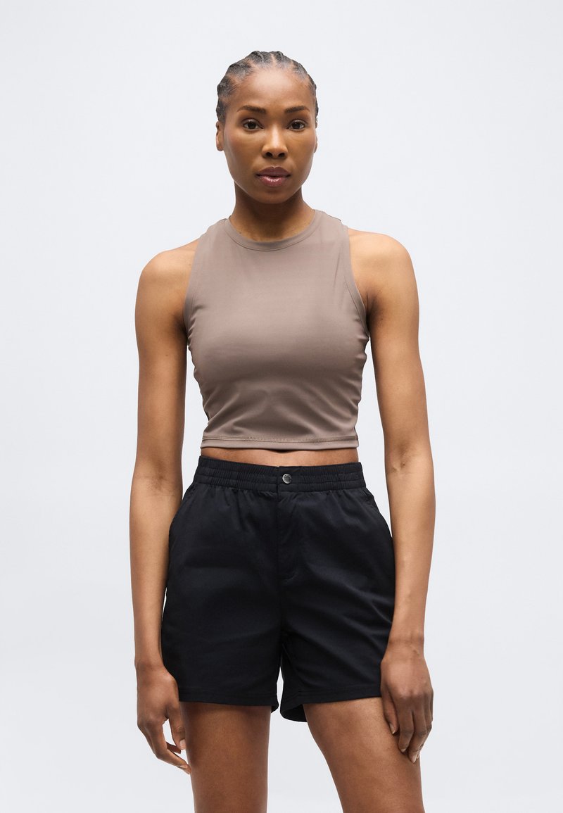 Γυναίκα με πλεξούδες, που φοράει μπλούζα τοπ χωρίς μανίκια σε ταουπέ απόχρωση και μαύρο ψηλόμεσο σορτς, στέκεται μπροστά σε απλό ανοιχτό φόντο.