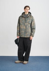 Kapucňový anorak s kamuflážným vzorom v zelených a hnedých odtieňoch, s prednou kapsou, skombinovaný s čiernymi širokými nohavicami a béžovou obuvou.