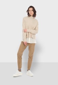 Beige geribbelde gebreide trui met hoge nek, gelaagd over een witte blouse, gecombineerd met lichtbruine broeken en witte sneakers.