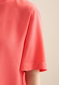 Blusa a maniche corte in corallo, realizzata in tessuto liscio. Presenta un design minimalista con un semplice scollo rotondo e maniche con risvolto.