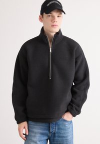Pulover din fleece negru cu guler înalt și închidere cu fermoar. Prezintă un material texturat, o croială confortabilă și un patch cu logo discret pe piept.