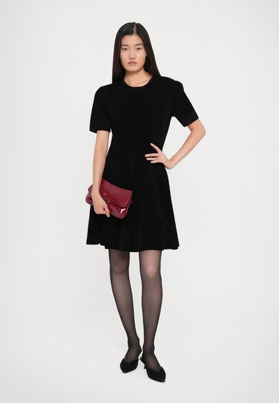 Woman standing wearing a black short-sleeve dress, black sheer tights, black heels, and holding a glossy red clutch bag.