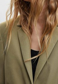 Blazer verde oliva con rever a punta, realizzato in un tessuto morbido. Abbinato a un top nero sottostante. Fili di capelli texturizzati visibili.