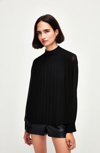 Blusa nera plissettata con colletto alto, maniche lunghe trasparenti e polsini a coste. Abbinata a pantaloni corti di pelle nera. Tessuto liscio ed elegante.