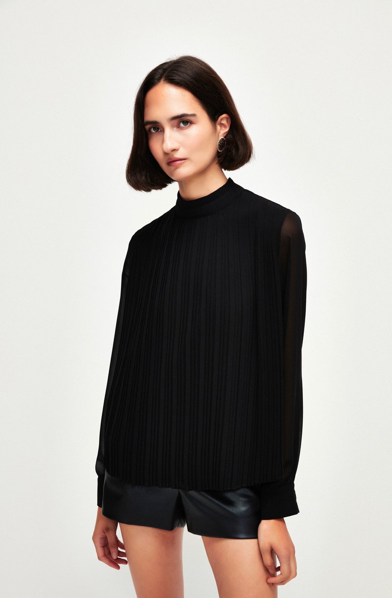 Sort plisseret bluse med høj hals, lange gennemsigtige ærmer og ribbede manchetter. Kombinationen med sorte lædershorts. Glat, elegant tekstur.