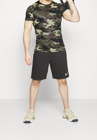 Kamouflerad t-shirt med kort ärm i mörkgröna och bruna mönster, matchat med svarta shorts och beigea träningsskor.