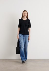 Chemise noire à manches courtes avec un col et des poignets à motifs, associée à un jean bleu clair ample et un sac noir. Fond simple.
