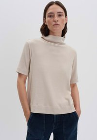Donna con capelli corti castani che indossa un maglione a collo alto beige a maniche corte e pantaloni blu scuro, in piedi con le mani in tasca su uno sfondo semplice.