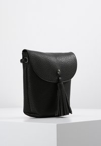 Svart läder crossbody-väska med en strukturerad yta, klaffstängning och fransdetalj. Kompakt kvadratisk form med en rund nitaraccent.
