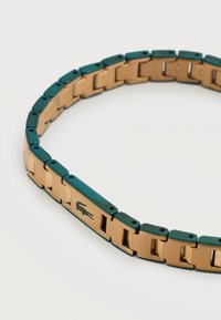 Βραχιόλι από χρυσό και πράσινο μέταλλο με ορθογώνιους κρίκους και μικρό λογότυπο κροκόδειλου σε έναν κρίκο, σε λευκό φόντο.