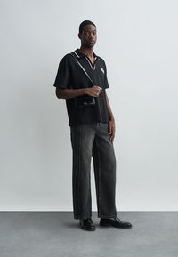 Μαύρο κοντομάνικο πουκάμισο με λευκό γιακά και λογότυπο, σε συνδυασμό με γκρίζα παντελόνια φαρδιάς γραμμής με πλαϊνές ραφές. Μαύρη τσάντα messenger και παπούτσια.