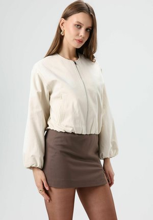 Beige bomberjack met een ritssluiting aan de voorkant, geplooide manchetten en een enkele zijzak, gecombineerd met een nauwsluitende bruine minirok.
