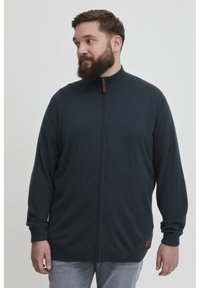 Tmavě modrá mikina na zip s vysokým límcem, dlouhými rukávy, žebrovaným spodním okrajem a manžetami, a malou koženou nášivkou na spodní straně.