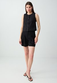 Femme aux cheveux bruns raides porte un top noir sans manches boutonné, un short noir taille haute et des sandales noires à talons, debout sur un sol blanc.