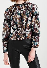 Kukkakuosisesti valkoinen pusero, jossa on monivärisiä kuvioita, pitkät hihat, kokoontuva etuosa ja mustat pitsiyksityiskohdat. Yhdistetty mustiin sukkahousuihin.