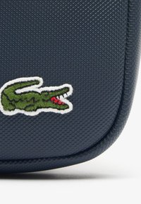 Mornarica teksturirana torba s zelenim aligatorskim logom u bijelom okviru. Ima glatku površinu i zaobljene rubove.