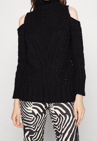 Pull noir à épaules dénudées en maille torsadée, associé à un pantalon noir et blanc à imprimé zèbre abstrait, porté par une personne debout.