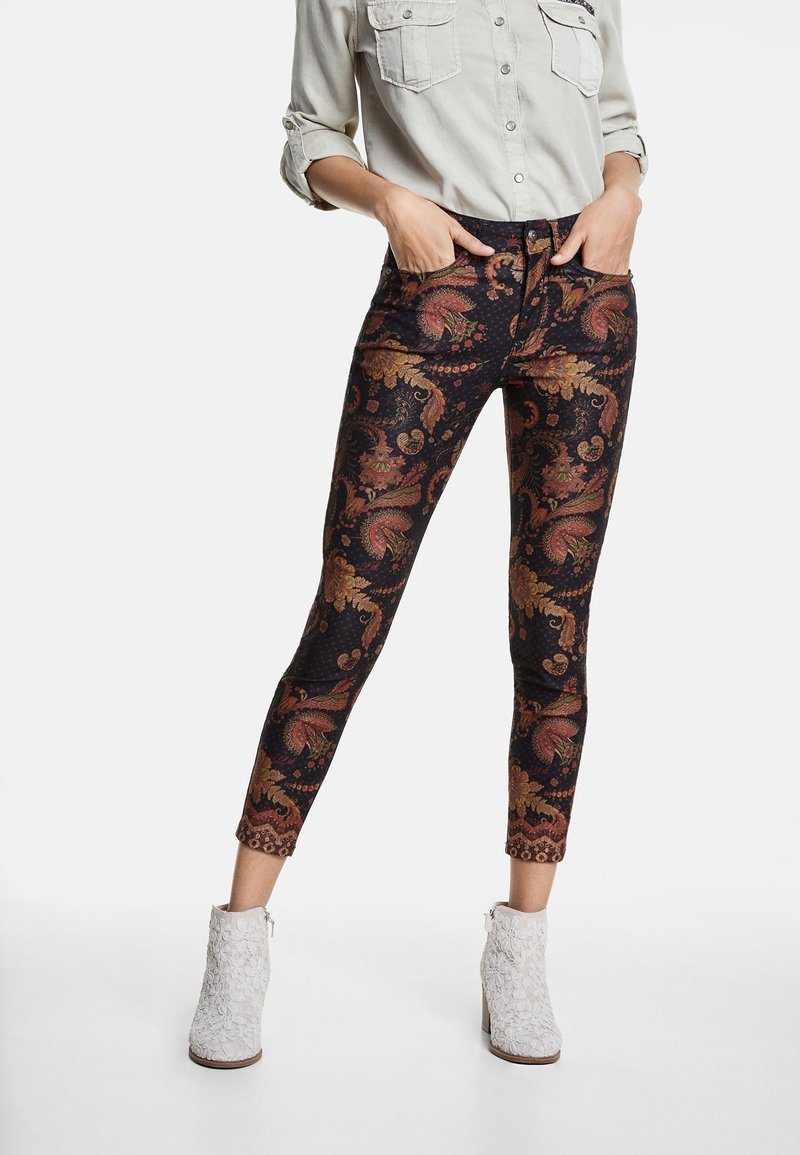 Jeans skinny en tissu noir avec un motif paisley doré et rouge. Dotés d'une taille haute et d'une coupe cheville. Portés avec des bottines blanches texturées.