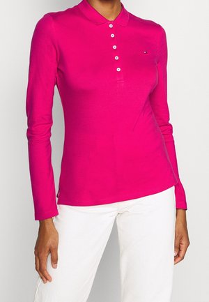 Femme portant un polo à manches longues rose vif avec des boutons et un pantalon blanc, debout devant un fond uni.