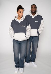 Hanorace în gri închis și gri deschis cu buzunare tip cangur. Ambele au șnururi și textul „MULTIPLY APPAREL” în alb. Blugi din denim purtați.