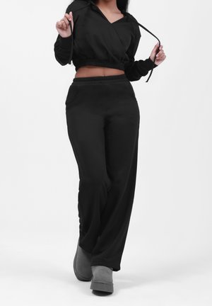 Kobieta ubraną w czarną krótką bluzę z kapturem i czarne szerokie spodnie spaceruje, trzymając sznurki od bluzy, ma na nogach szare botki na białym tle.