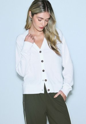 Nuori nainen, jolla on pitkät vaaleat hiukset, valkoinen napitettava neuletakki ja tummat housut. Hän seisoo ja katsoo alaspäin, käsi taskussa.