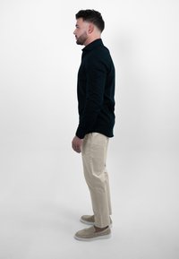 Donkerblauwe lange mouwen shirt, beige chinos, lichtbruine instapschoenen. Zijaanzicht toont een aangesloten ontwerp en een gladde stoftextuur. Effen achtergrond.