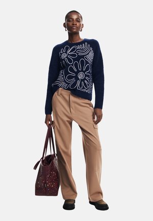 Pull en mailles bleu marine avec des motifs floraux blancs, associé à un pantalon taille haute beige et un sac fourre-tout perforé bordeaux. Chaussures noires.