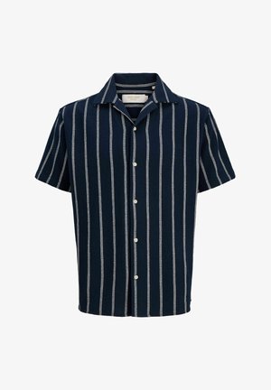 Adolescente con indosso una maglietta a maniche corte a righe verticali bianche e nere e pantaloncini abbinati, con scarpe da ginnastica bianche e un cappellino blu.