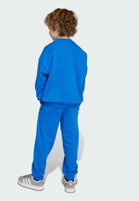 Sweatshirt en coton bleu et pantalon assorti avec des poignets élastiques, présentant une coupe décontractée. Associé à des baskets grises avec des rayures blanches.