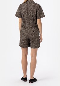 Luipaardprint jumpsuit in beige en zwart. Korte mouwen, elastische taille, en shorts. Voorzien van een logopatch op de achterkant. Zwarte schoenen.
