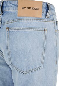 Lyseblå denimjeans med baklommer, synlige sømmer og en lærlabel med teksten "2Y STUDIOS" festet til midjebåndet.