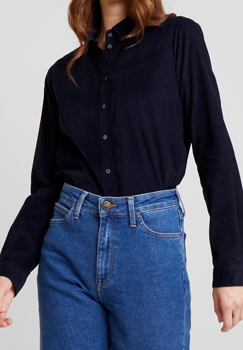 Femme portant une chemise noire à manches longues boutonnée, rentrée dans un jean en denim bleu taille haute avec un bouton visible de la marque Lee.